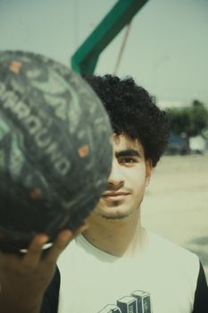 A young man holding a basketball confidently on an outdoor court, showcasing style and focus.