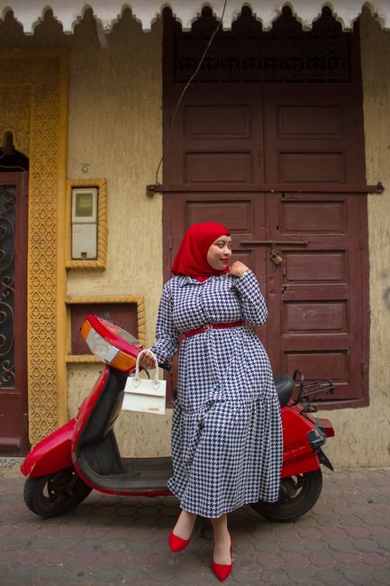 Stylish woman in a patterned dress and hijab standing by a red scooter on a city street.
