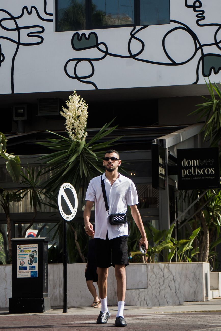 Stylish young man in sunglasses and black shorts walking on a city street with artistic wall art in the background.