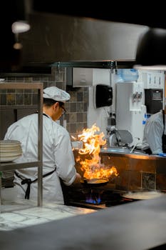 A chef skillfully handles a flaming pan in a commercial kitchen, showcasing culinary expertise.