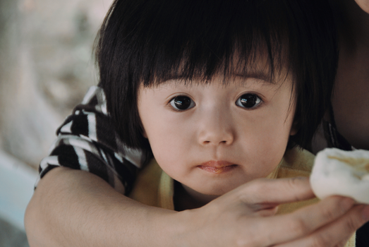 Cute Asian child in parent's arms, holding a snack, with an expressive gaze.