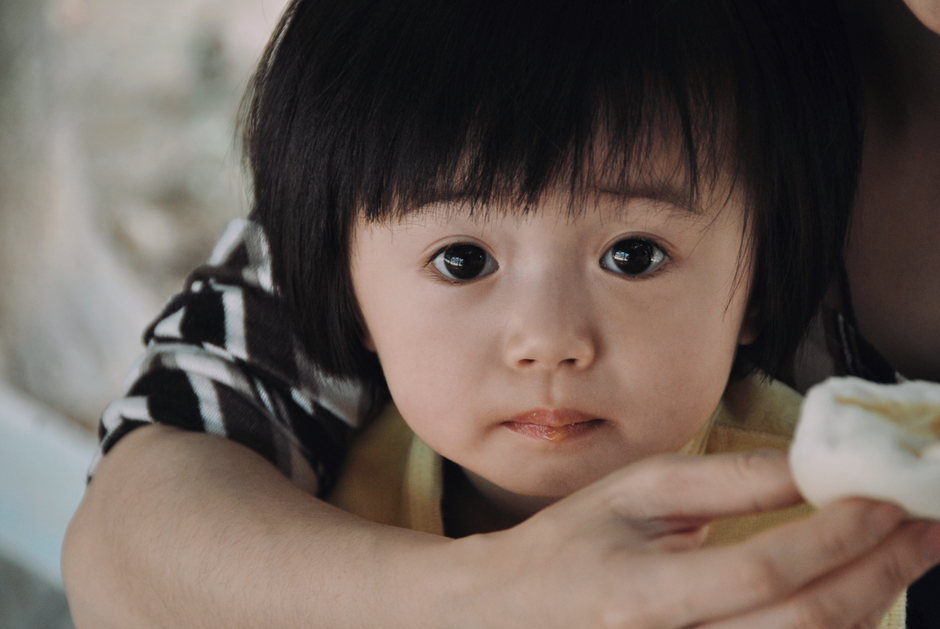 Cute Asian child in parent's arms, holding a snack, with an expressive gaze.