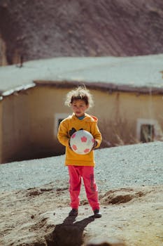 A young child holding a soccer ball, standing outdoors in a sunny rural area.