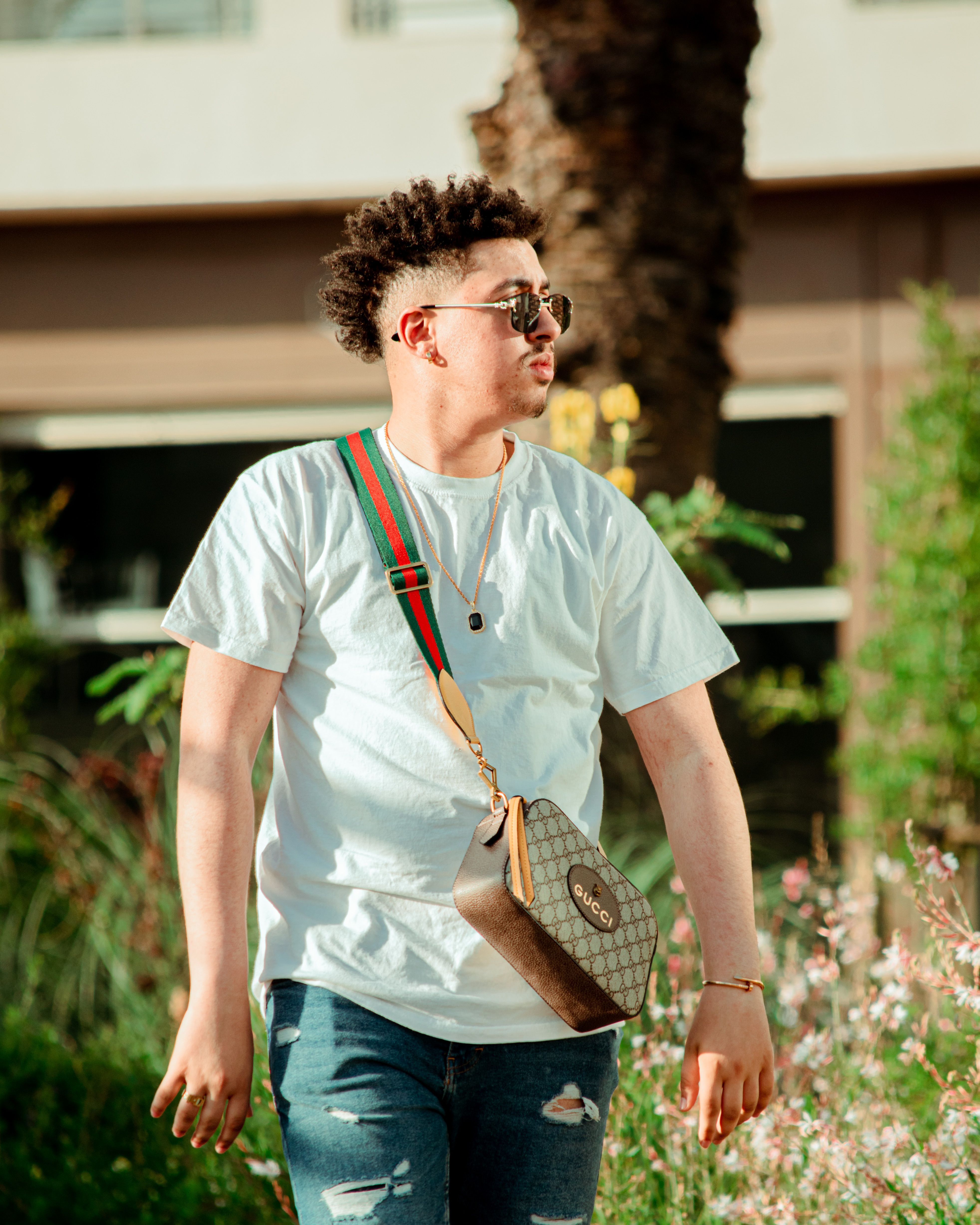 Stylish young man with sunglasses and crossbody bag walking outdoors on a sunny day.
