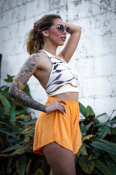 Fashionable woman with tattoos poses in sunny outdoor setting with sunglasses.