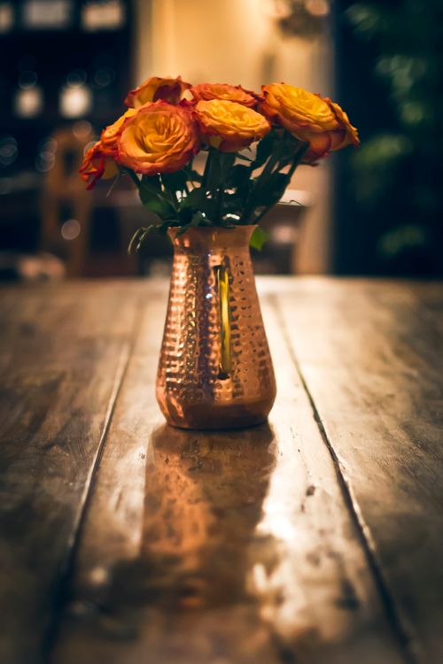 Free A handcrafted copper vase filled with colorful roses sits elegantly on a wooden table in warm, indoor lighting. Stock Photo