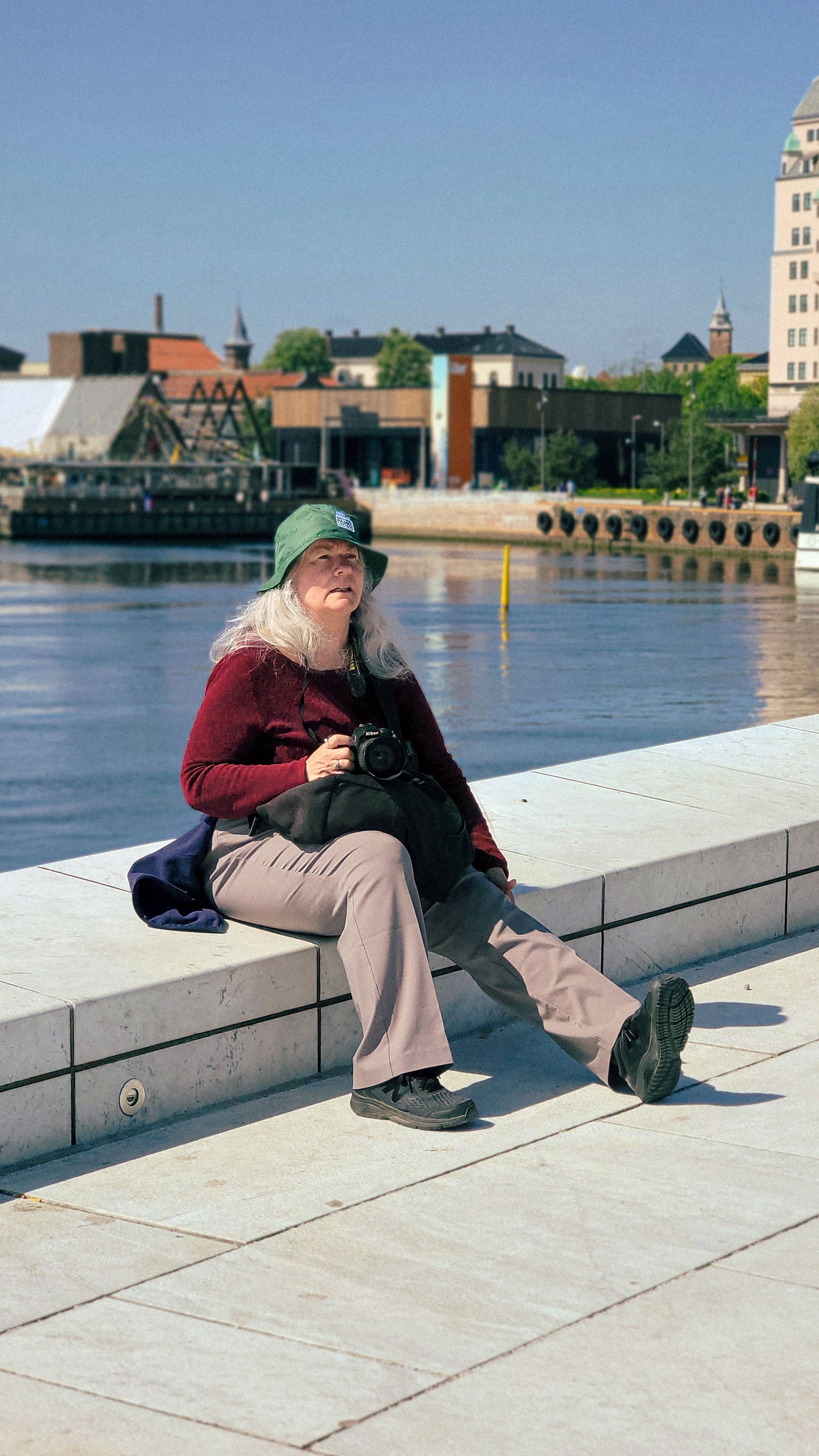 A senior woman photographer relaxing at Oslo's scenic waterfront, capturing vibrant urban views.