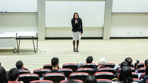 Female speaker presenting to an audience in a modern auditorium setting.