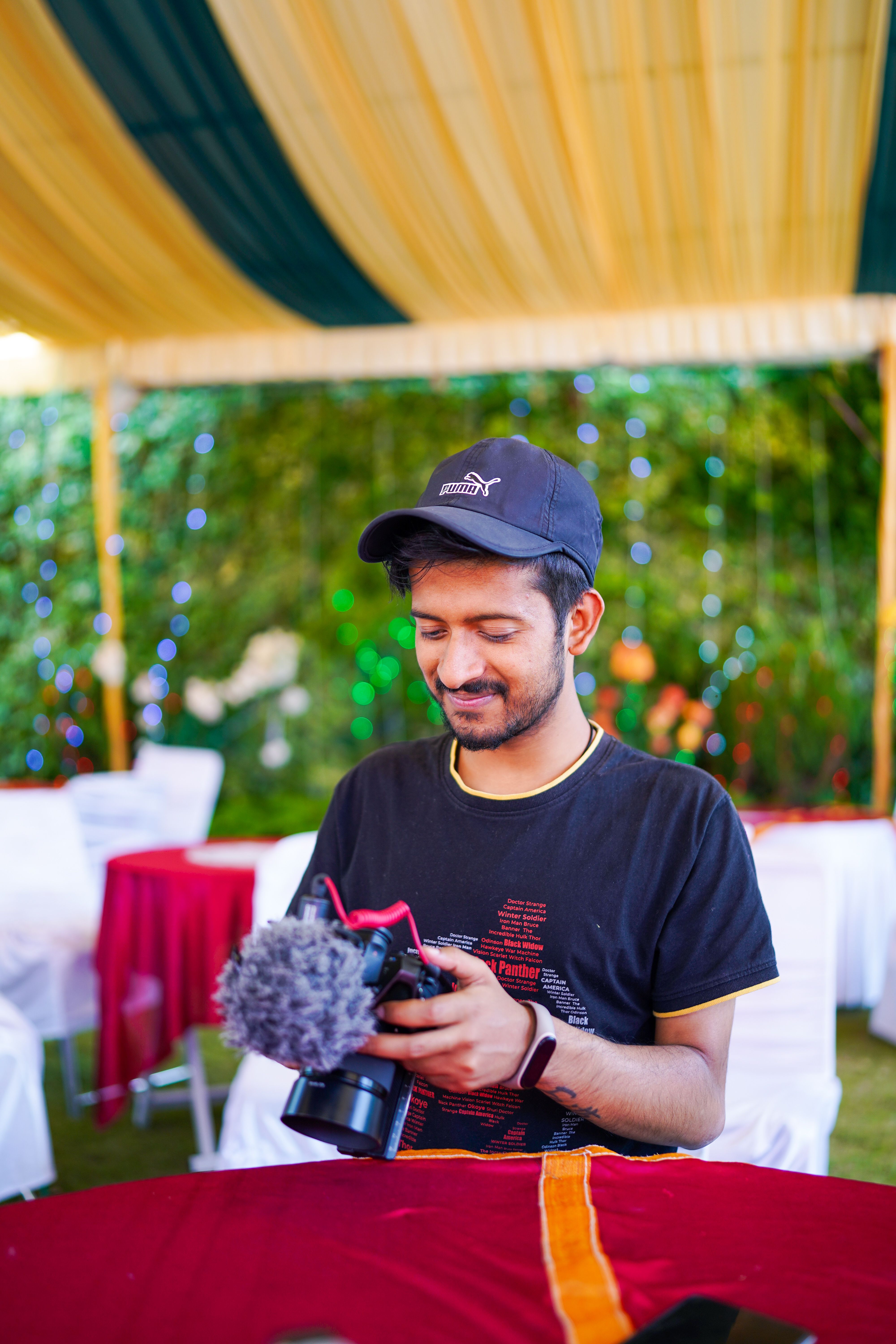 A young man in a black t-shirt holding a camera, smiling outdoors in Srinagar.