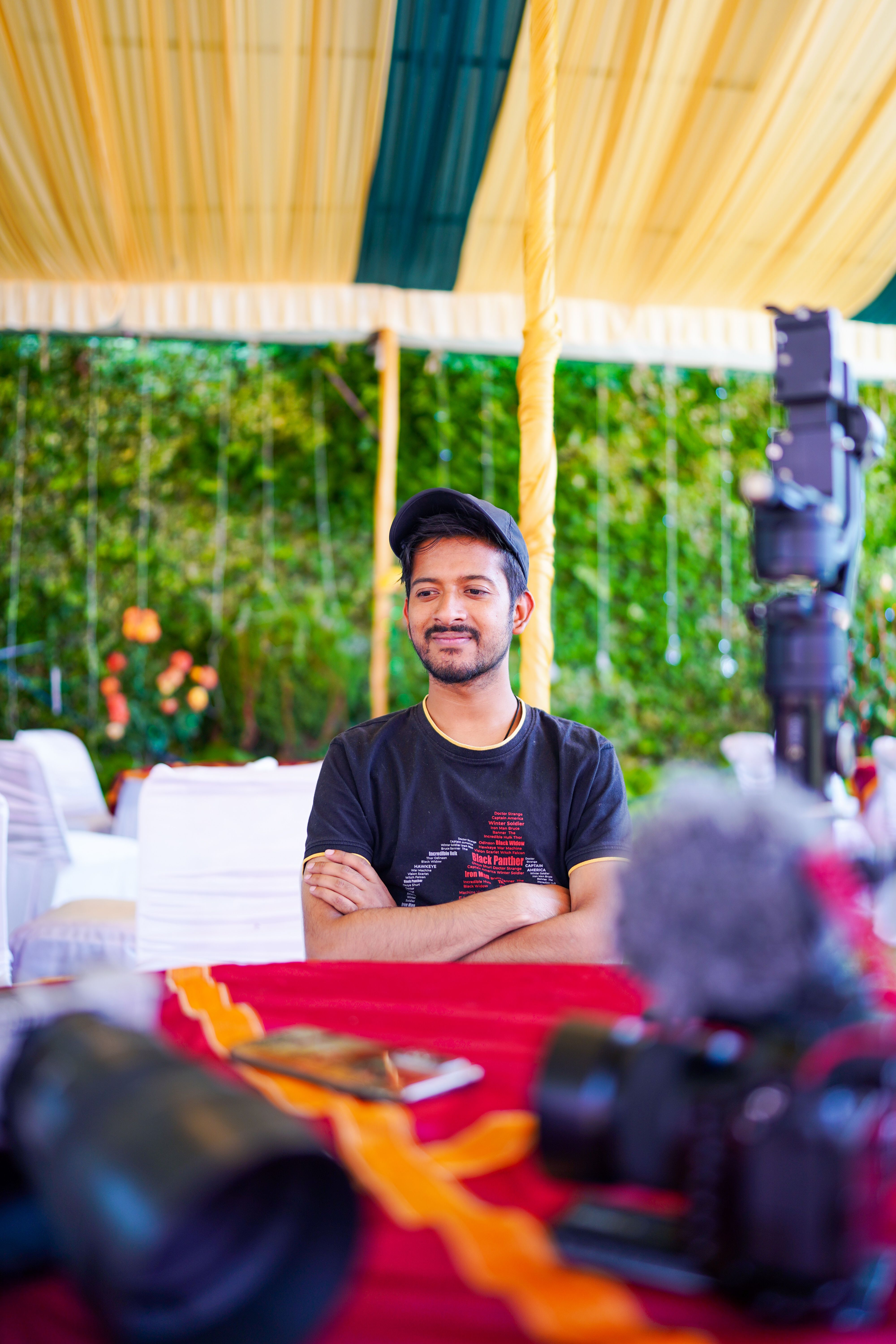 Smiling man in a cap and t-shirt, sitting at a table with photography equipment in an outdoor setting.
