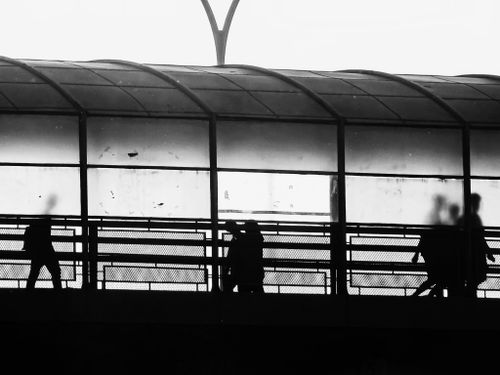 Free A black and white shot capturing silhouettes of people walking inside a glass tunnel in an urban setting. Stock Photo