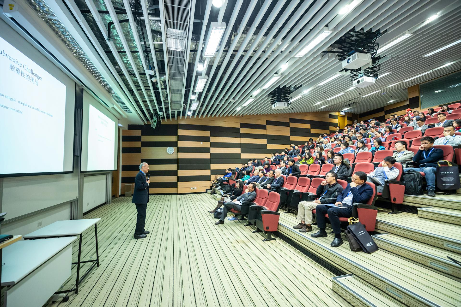 A speaker delivers a presentation to an attentive audience in a modern auditorium setting.