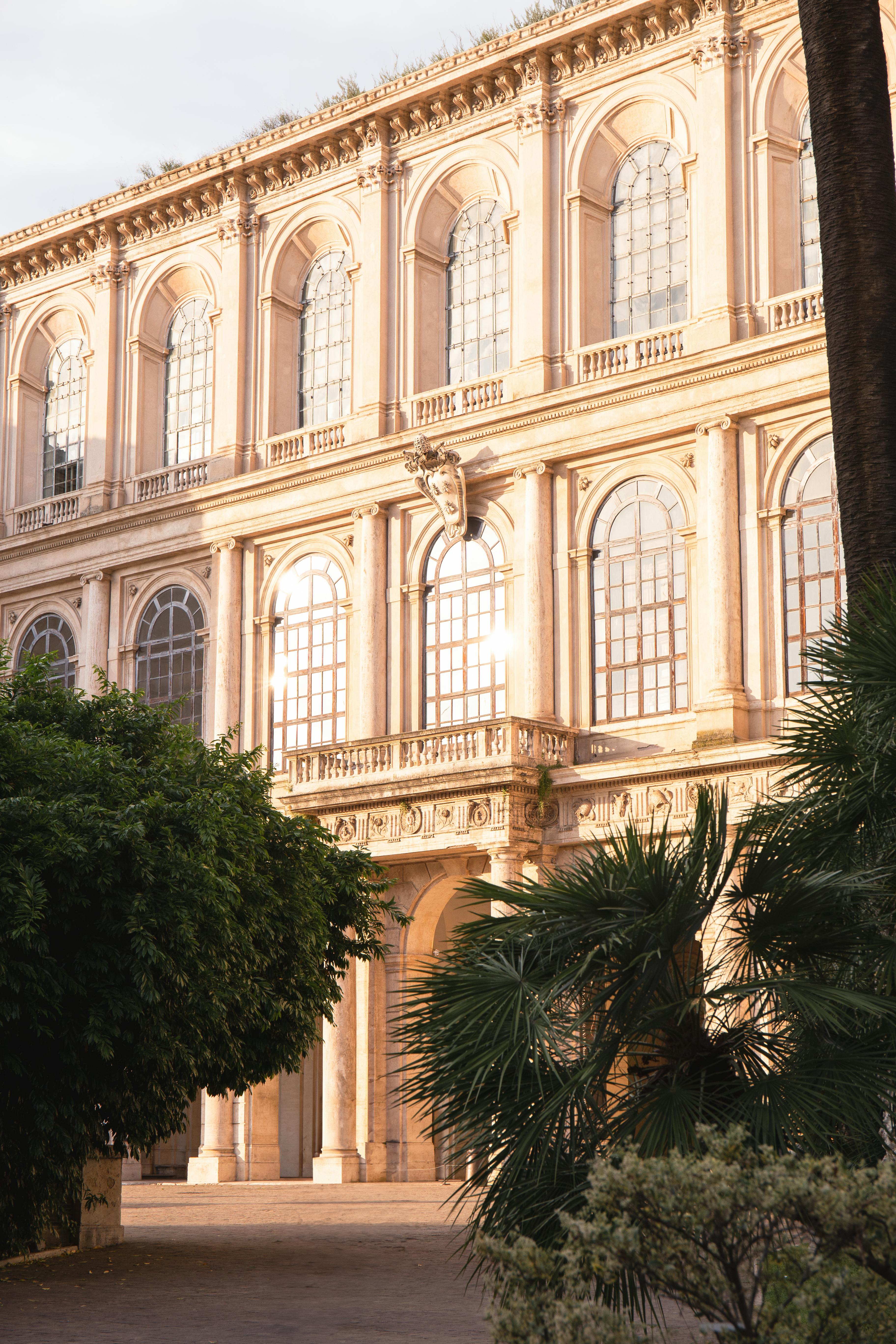 A Renaissance-style building facade in Rome, featuring tall windows and sunlight filtering through palms.