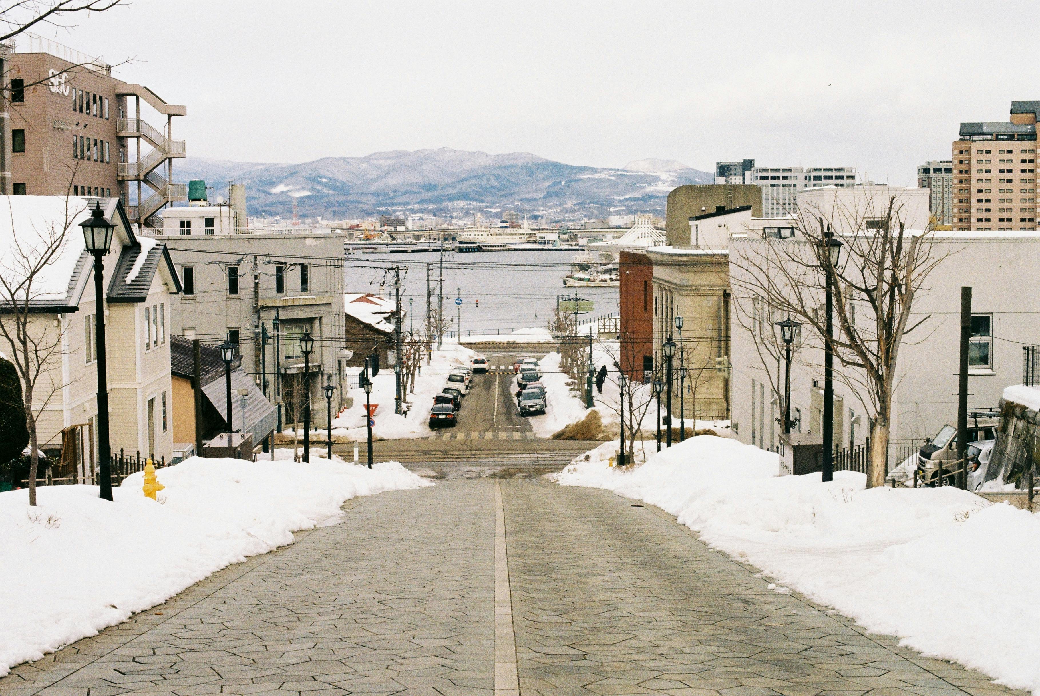 Snow-covered urban street leading to a bay, surrounded by city buildings and distant mountains.