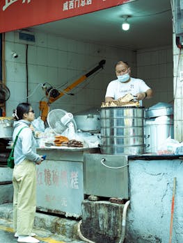 A street food vendor wearing a mask serves a customer in an urban market setting, showcasing steaming cuisine.