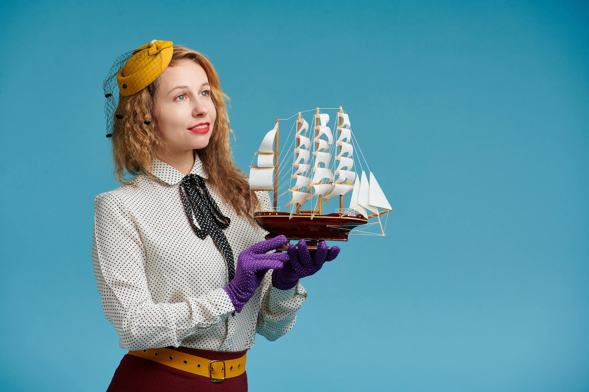 Vintage-style portrait of a woman holding a model sailing ship against a blue studio background.