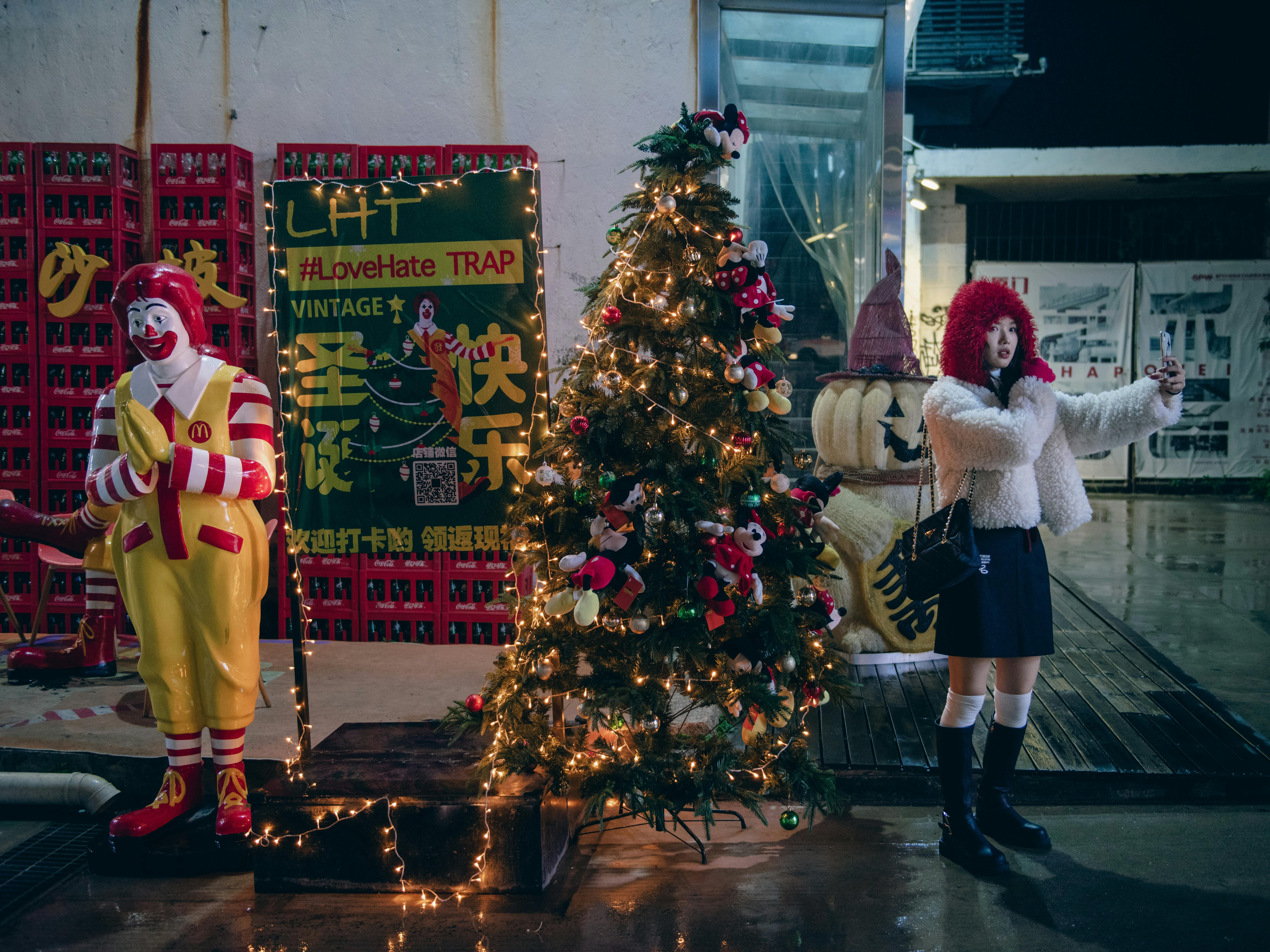 Woman taking a selfie near a vintage Christmas setup with iconic clown figure on city sidewalk at night.