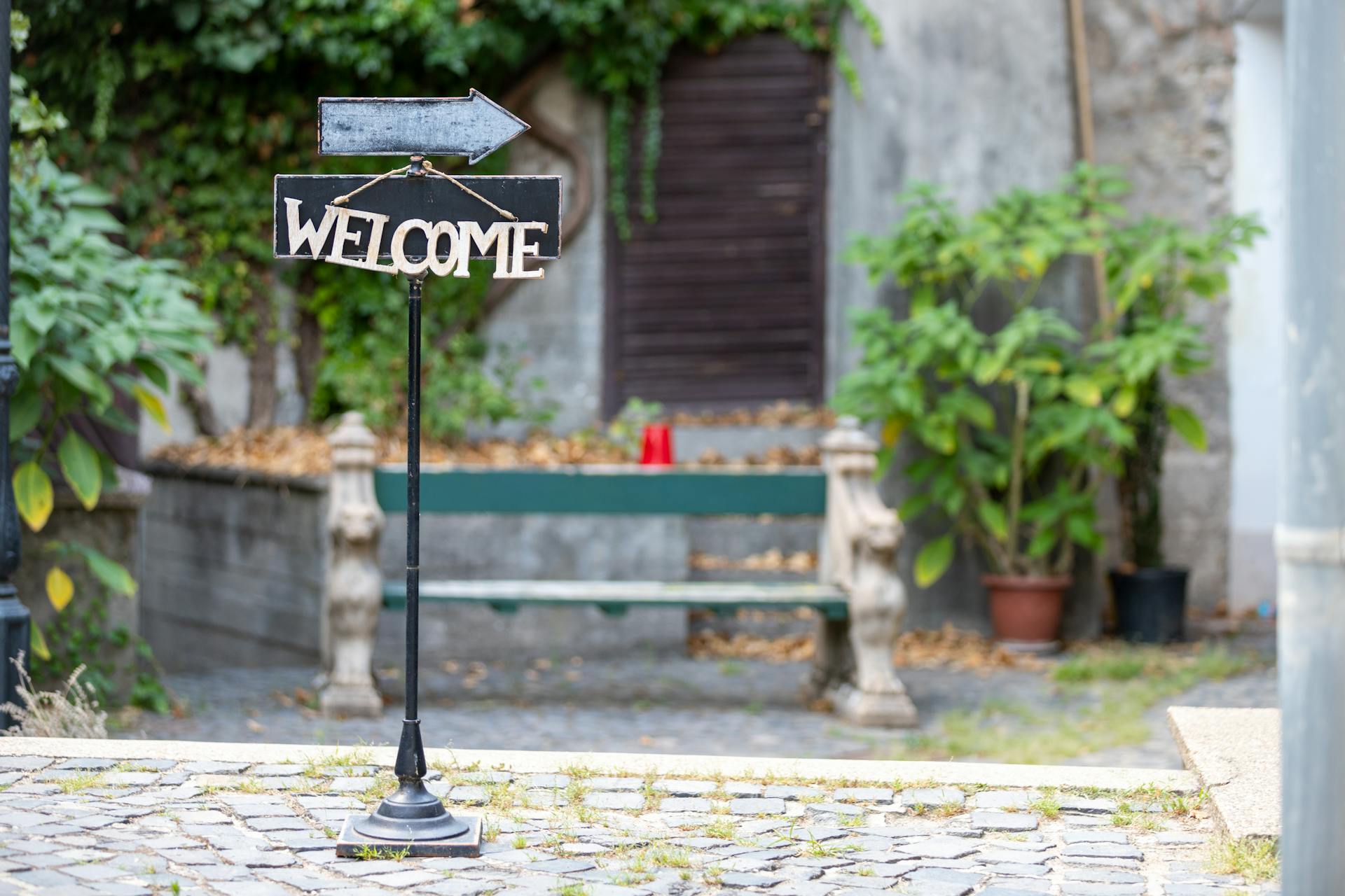 A welcoming sign with an arrow on a cobblestone walkway in a rustic urban setting.