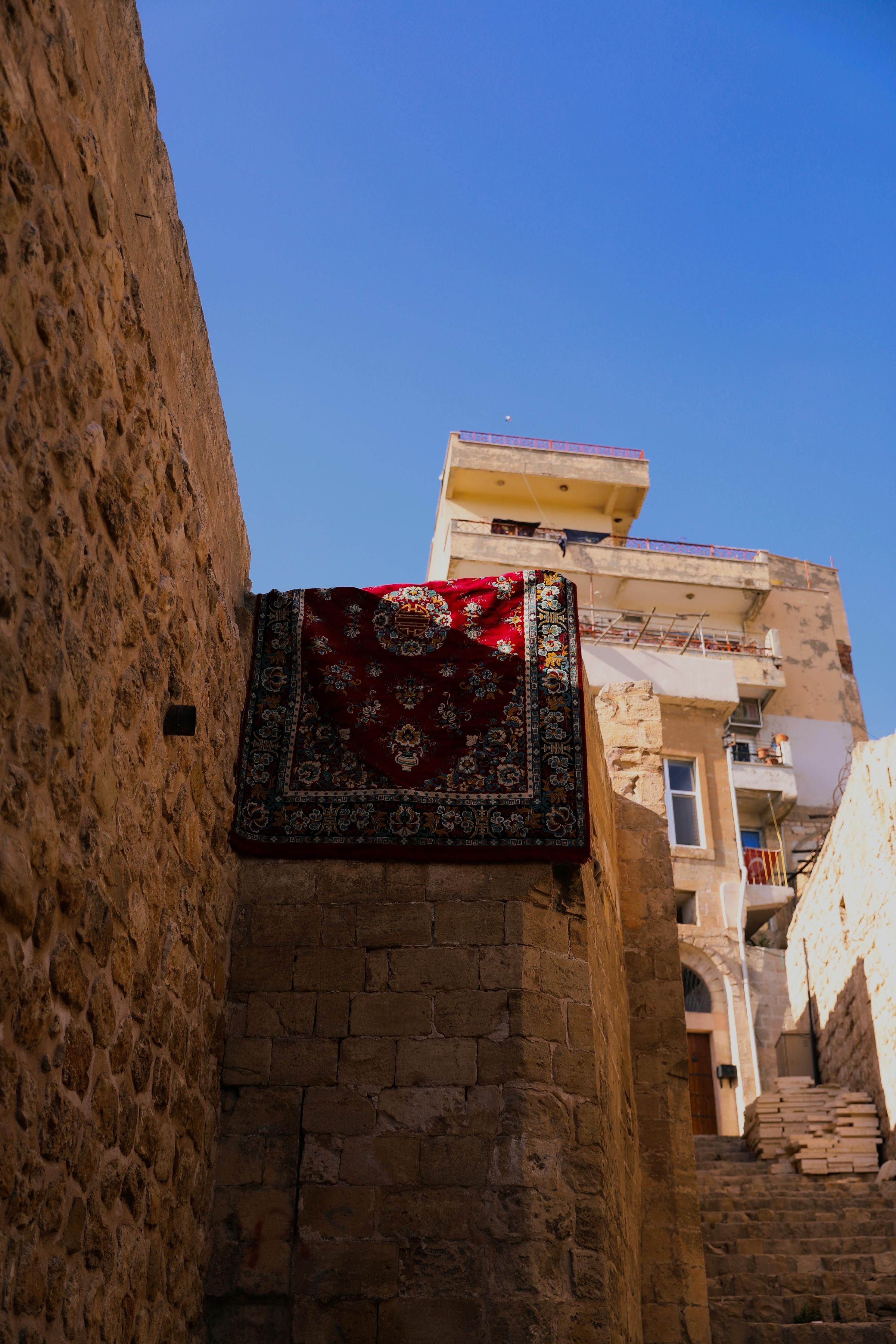 Red patterned rug draped over stone wall in narrow alley of historic town with blue sky backdrop.