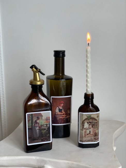 Three artistic bottles with vintage labels and a lit candle in a minimalist setting.