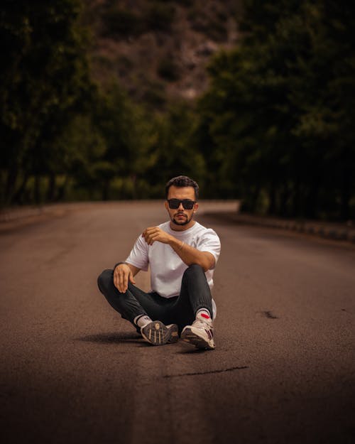 Man in a Casual Outfit and Sunglasses Sitting in the Middle of a Road