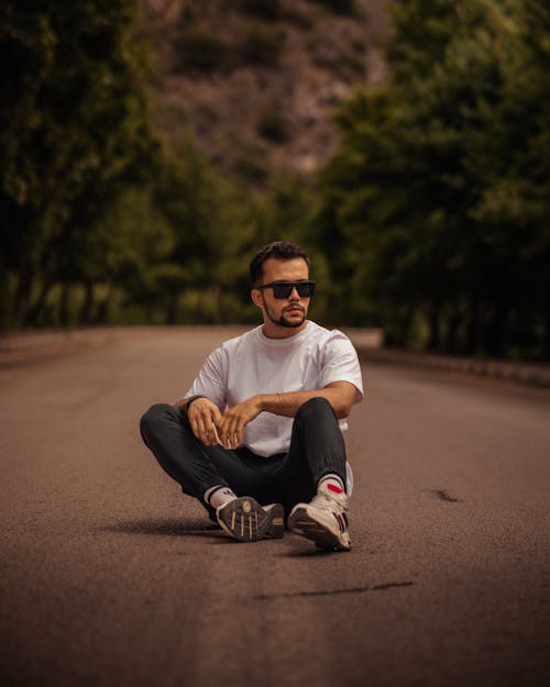 Man in a Casual Outfit and Sunglasses Sitting in the Middle of a Road