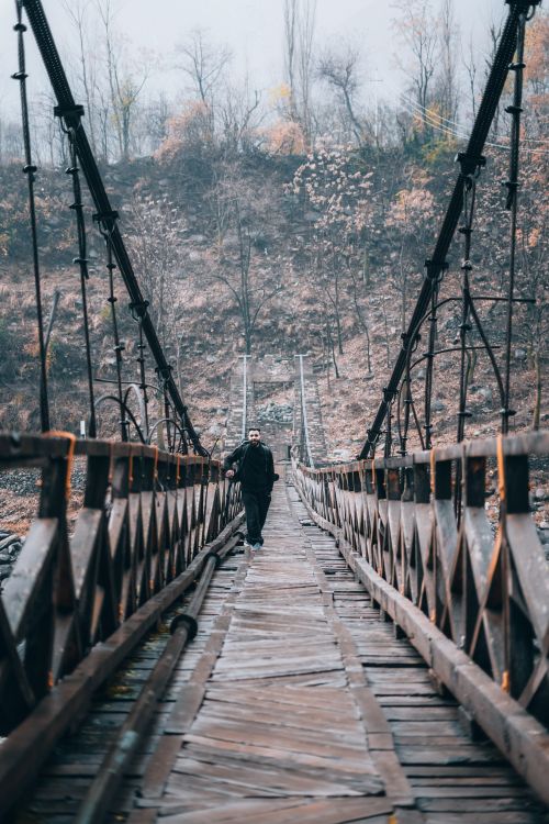 Free A solitary man walks across a rustic wooden suspension bridge surrounded by autumn foliage. Stock Photo