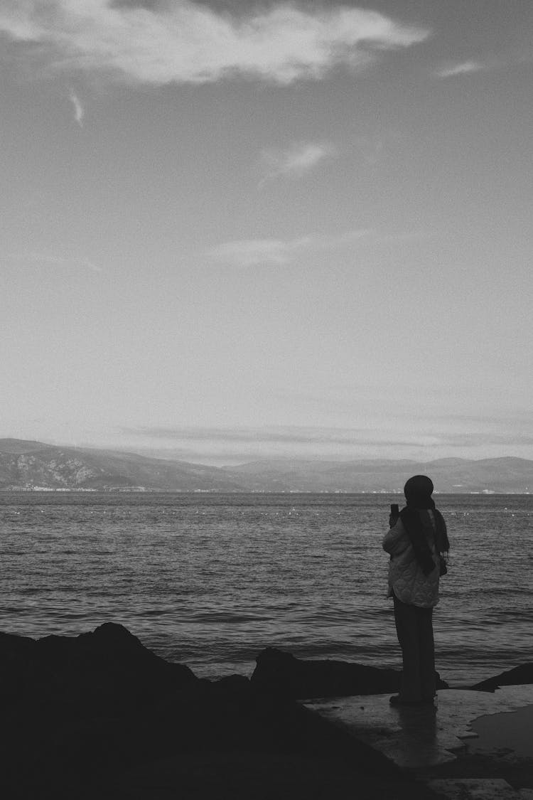 Black And White Photo Of A Woman In Headscarf Standing On A Seashore And Looking At Sea