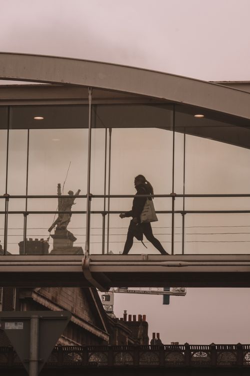 Free Woman walking on a modern glass footbridge in an urban cityscape, featuring a sculpture in the background. Stock Photo