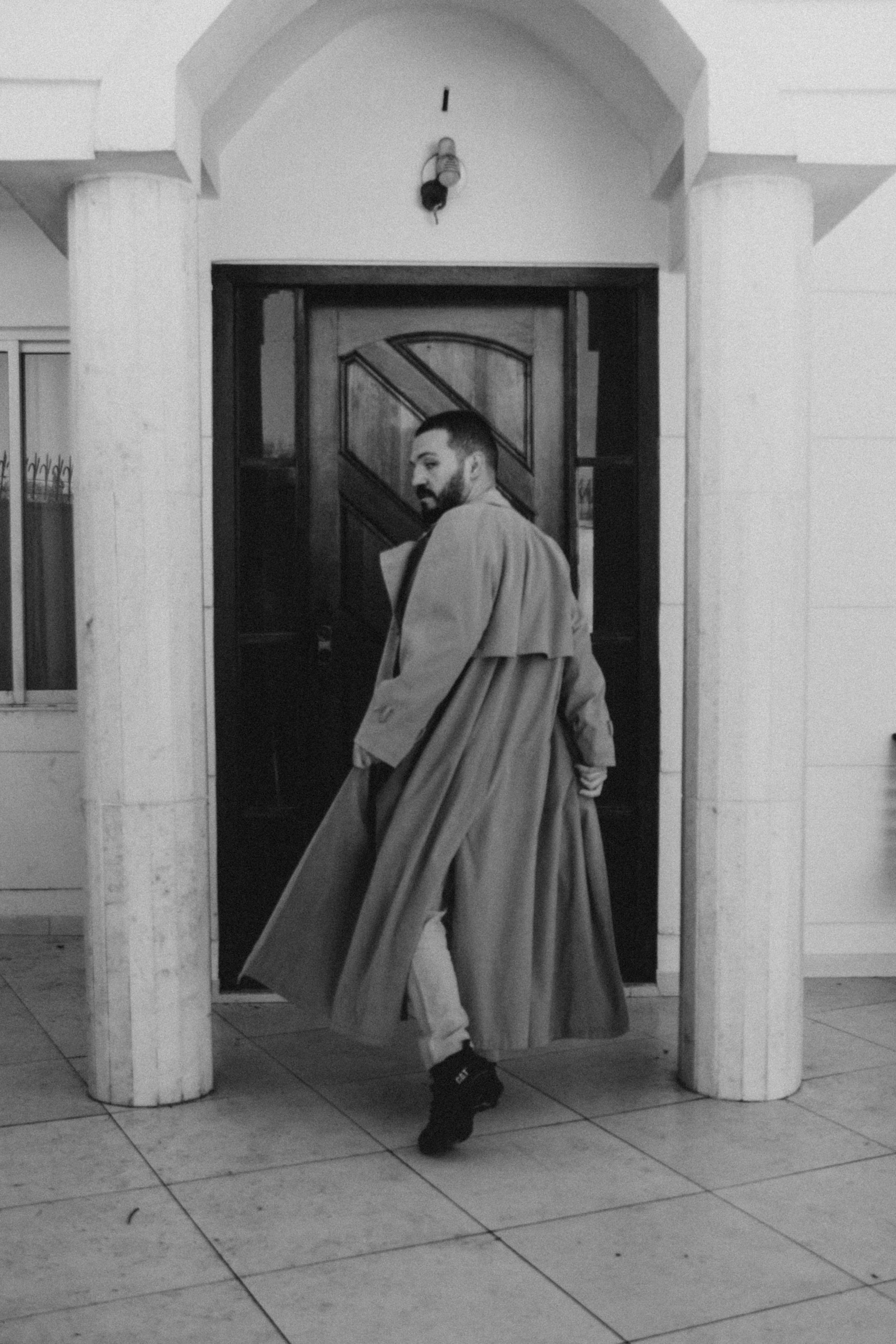 Black and white photo of a man in a trench coat turning near a classic doorway, embodying modern fashion.