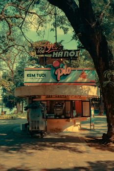 Vintage-style ice cream stand under trees with neon sign in Hanoi park during summer day.