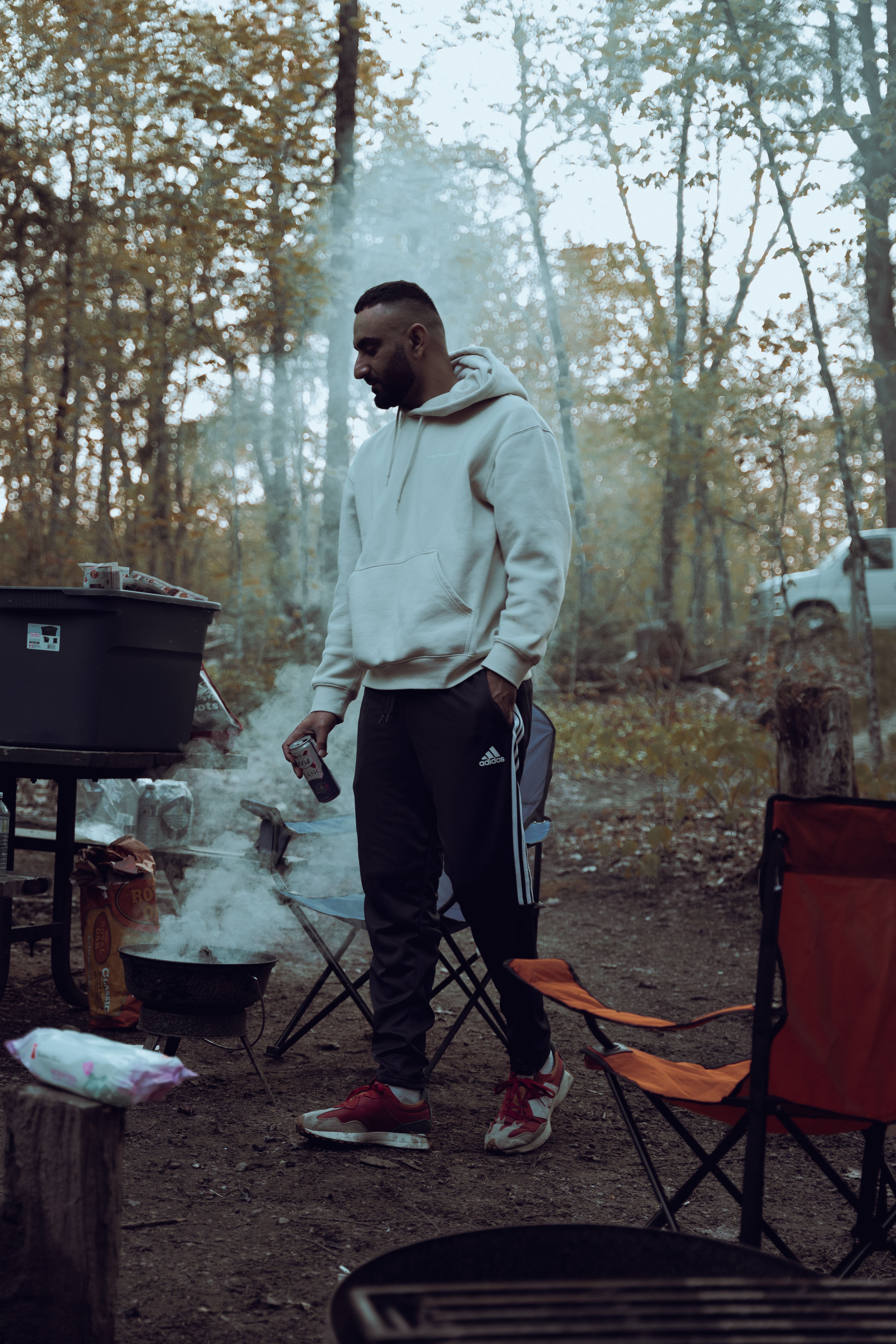 A man in casual attire stands beside a barbecue grill at a forest campsite, holding a beverage.
