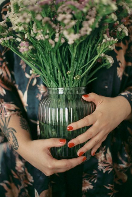 A woman with red nails holds a green glass vase filled with colorful flowers.