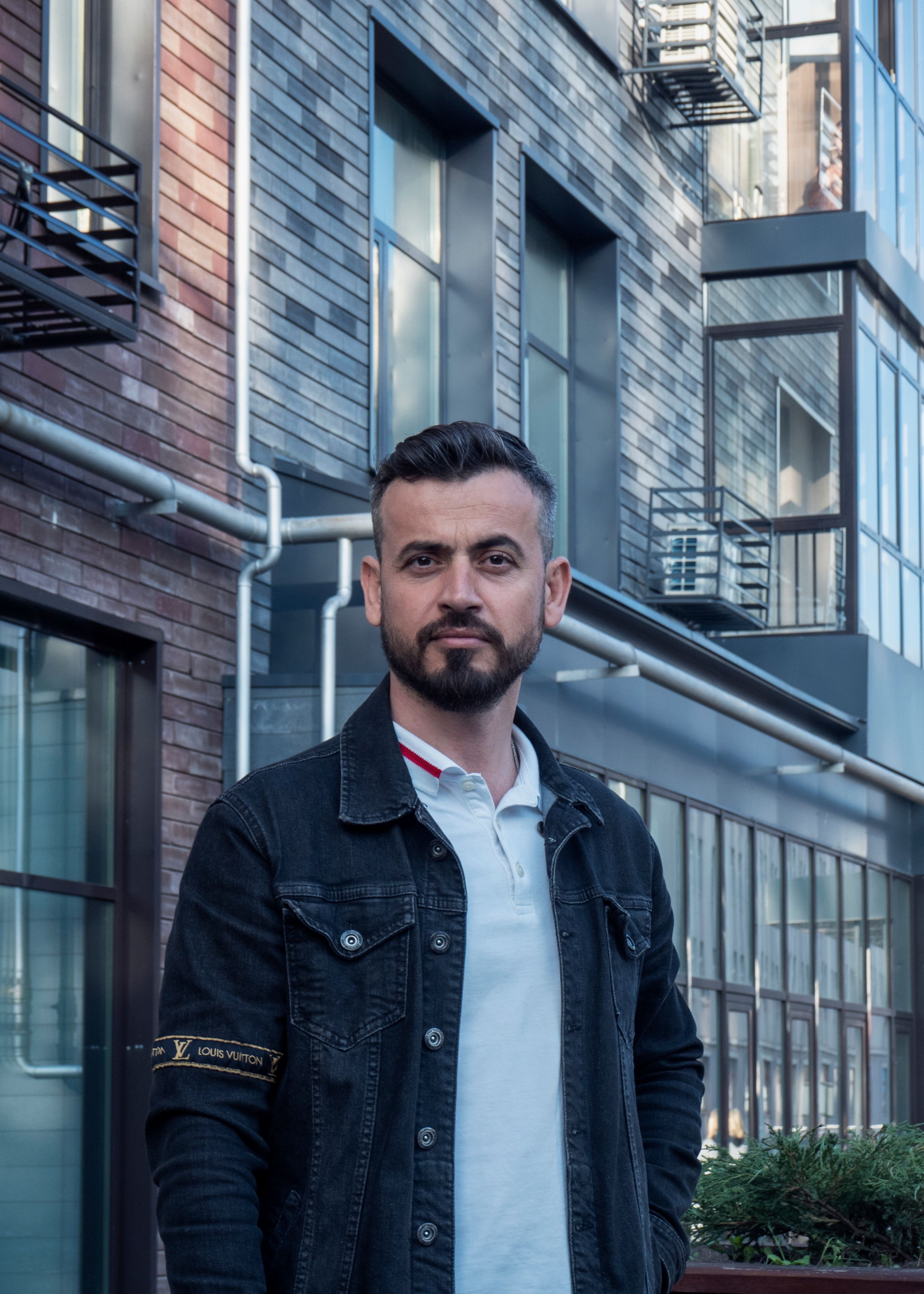 Portrait of a bearded man in a denim jacket set against a modern urban building backdrop.