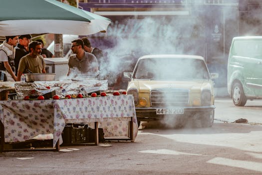 A yellow vintage Mercedes parked by a bustling urban food street with smoke enhancing the vintage ambiance.