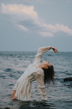 Woman in a white dress striking a dramatic pose by the sea, showcasing elegance and natural beauty.