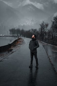A man looks up while standing on a wet road surrounded by a foggy, winter landscape.