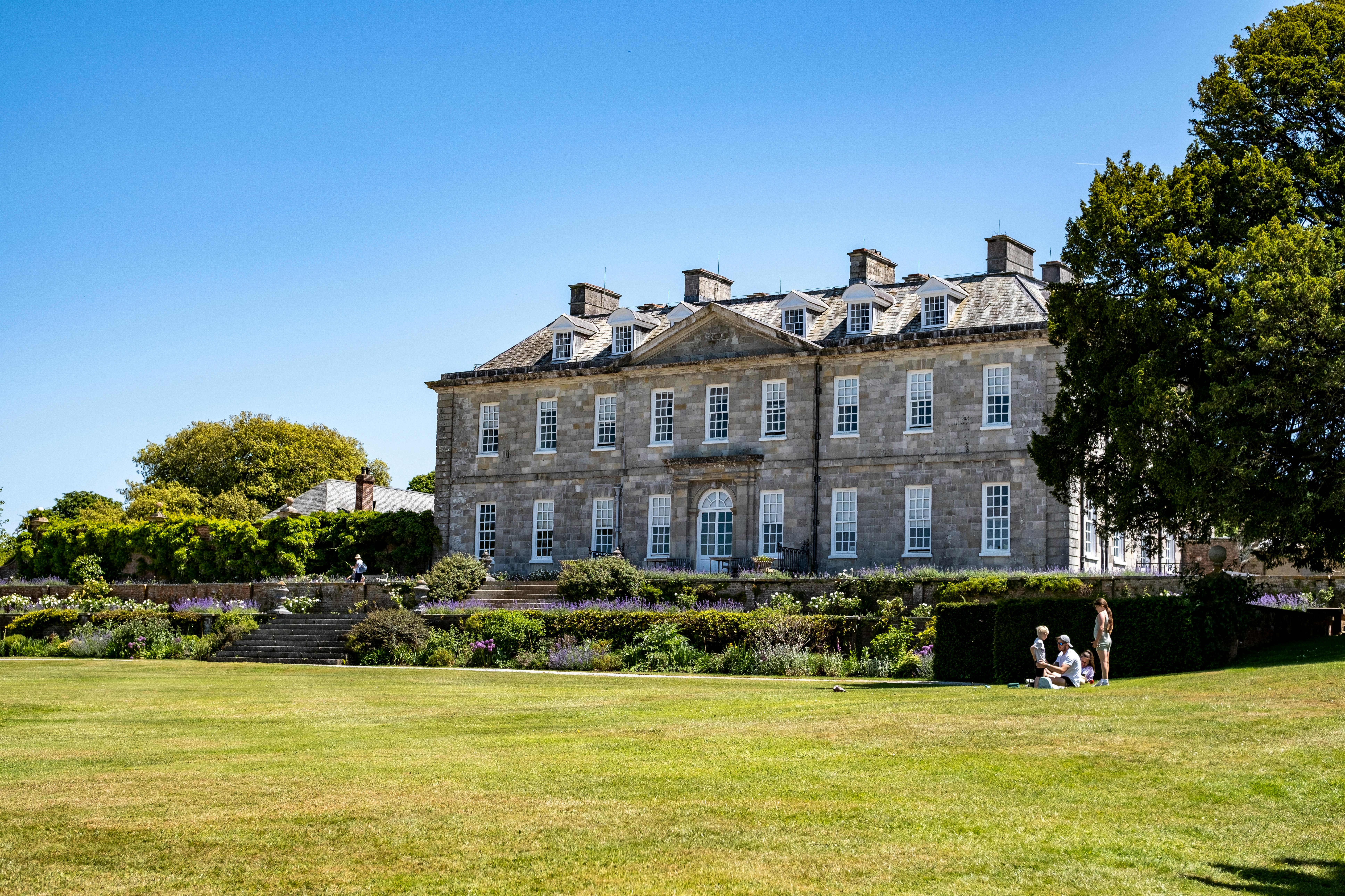 A historic stone mansion surrounded by lush gardens on a sunny day with people relaxing on the lawn.