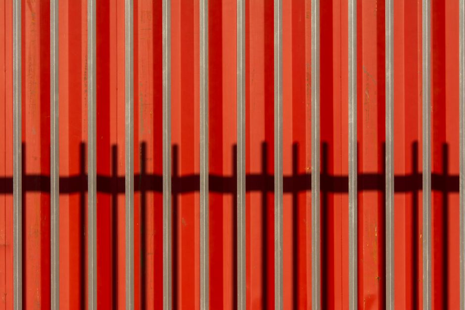 Close-up of sunlit red sheet metal with vertical bars casting shadows creating a striking pattern.