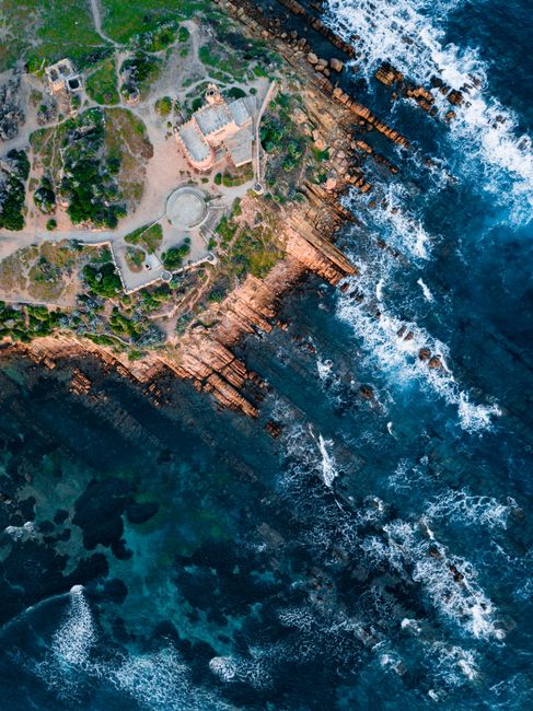 Drone shot of rocky coastline with ancient ruins and turbulent ocean waves.