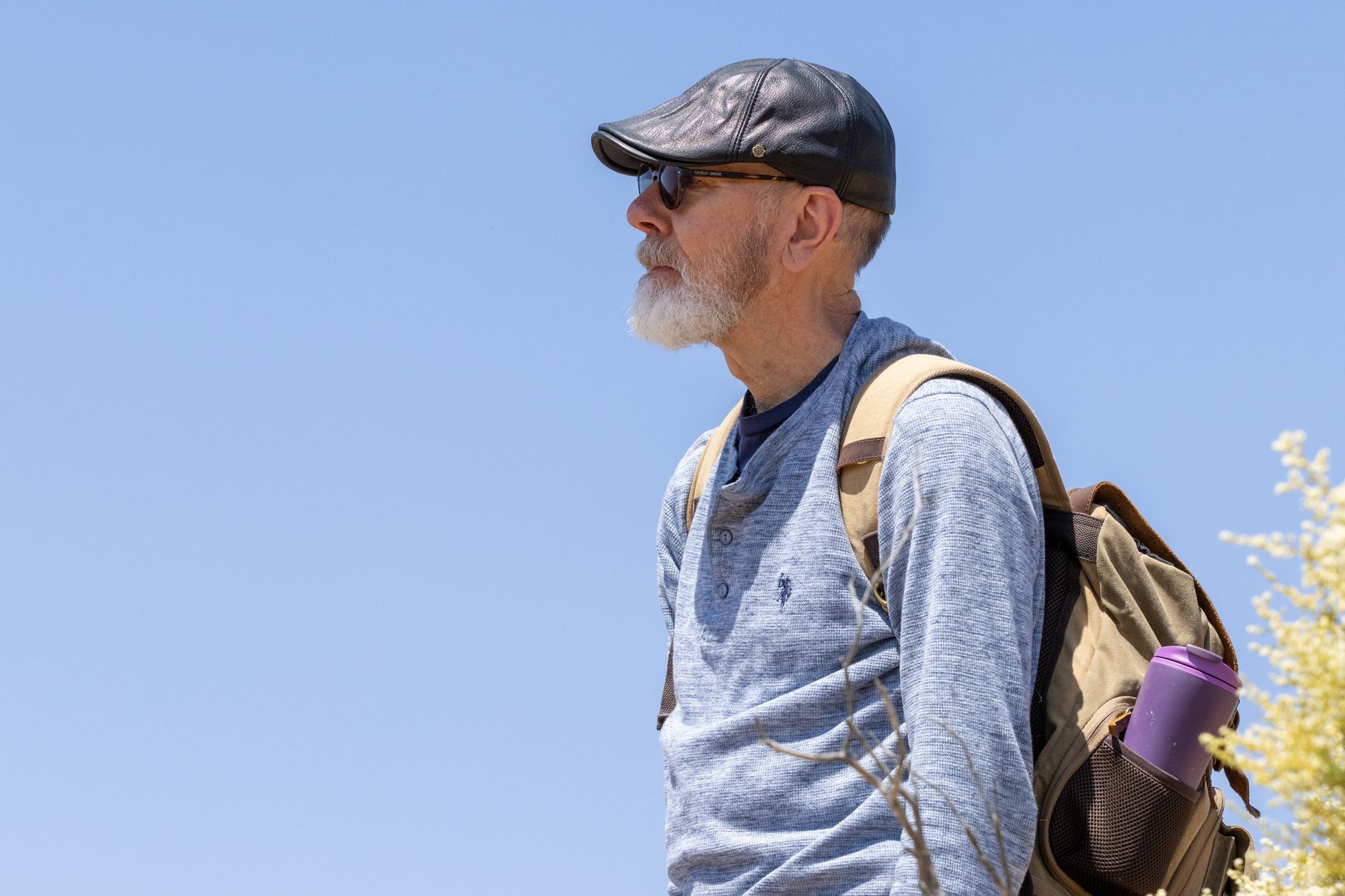 Portrait of an elderly man with a backpack hiking outdoors under a clear blue sky.