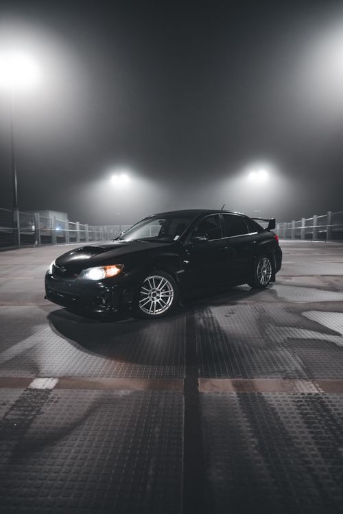 Free A stylish black sports car illuminated under night lights in a foggy parking lot. Stock Photo