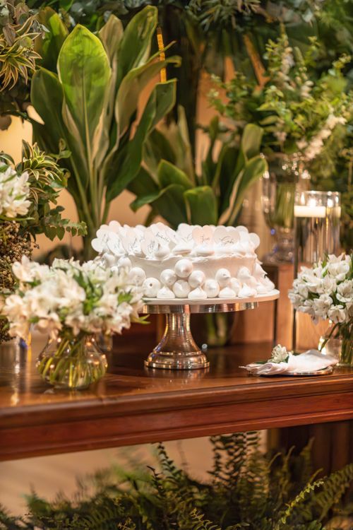 Free Elegant white cake on a stand surrounded by lush green foliage and white floral arrangements on a wooden table. Stock Photo