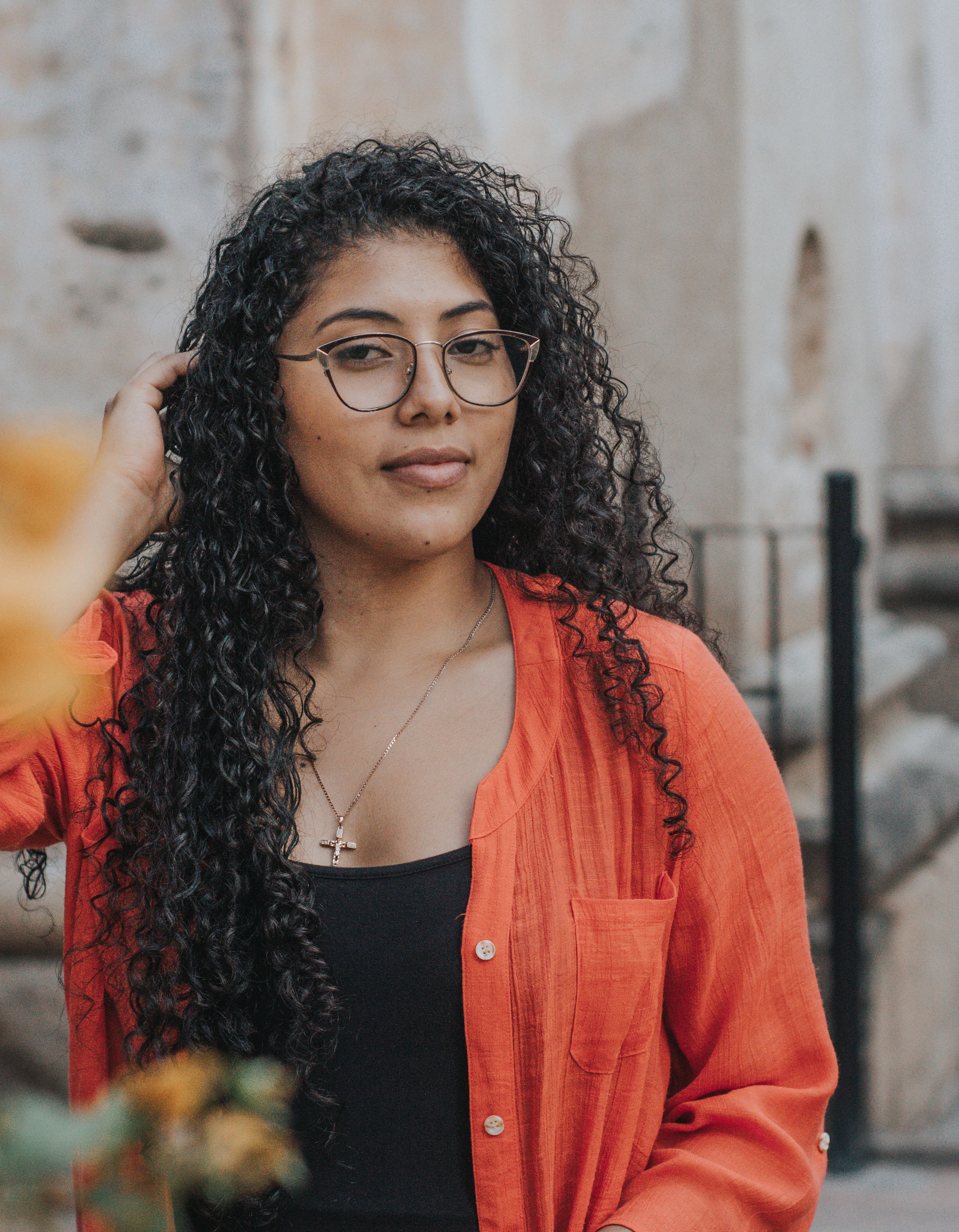 Portrait of a stylish woman with curly hair and glasses in a vibrant orange shirt outdoors.