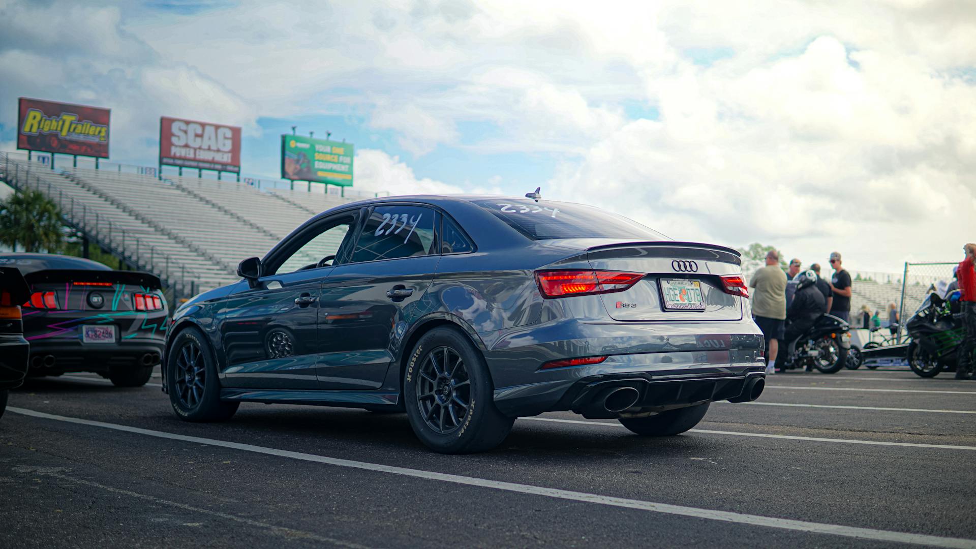 Audi S3 sports car at a racetrack gathering with grandstands and people in the background.