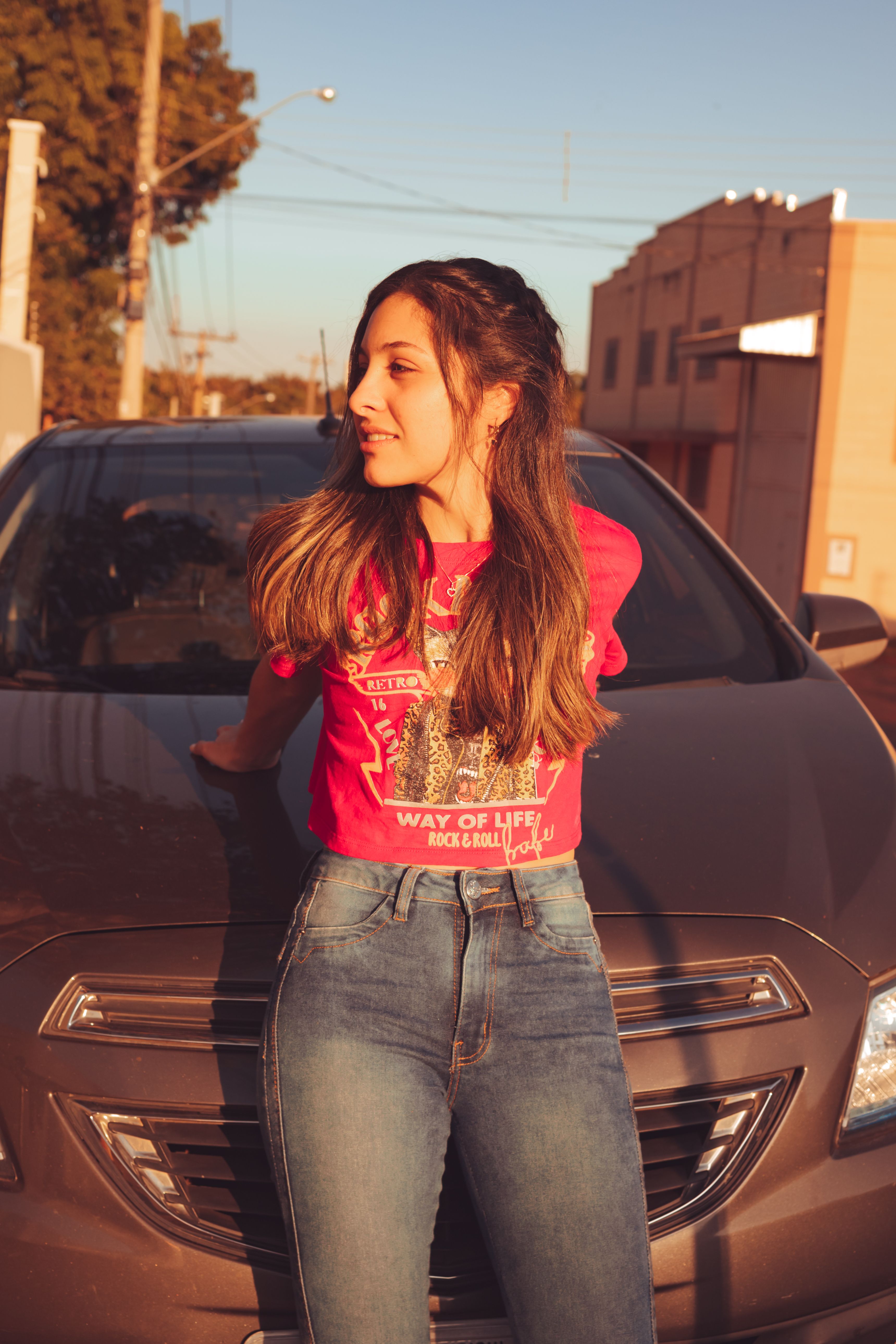A young woman in casual attire leans on a car hood during a sunny summer day.