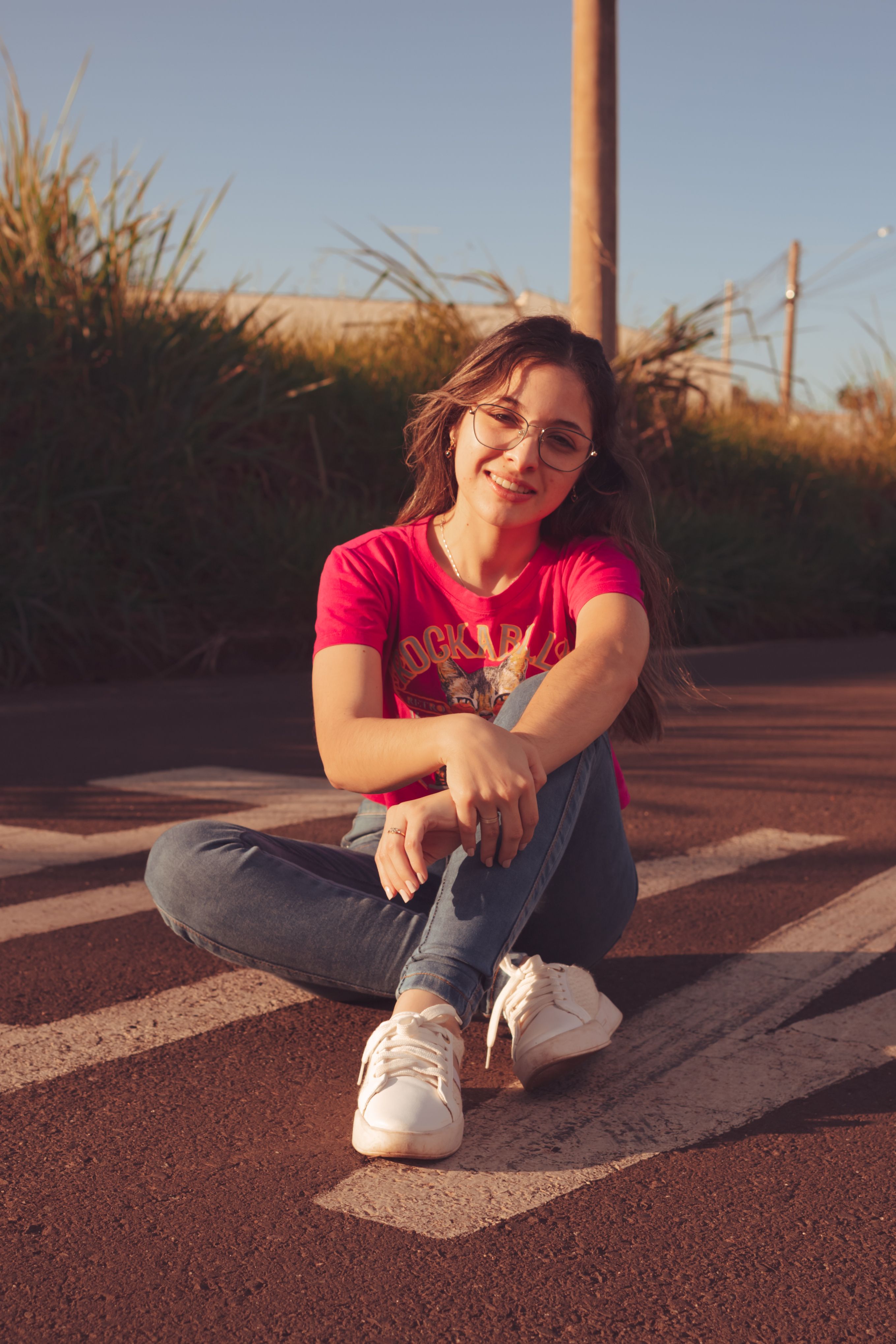 Young woman in casual outfit sits on the street, smiling on a sunny day.