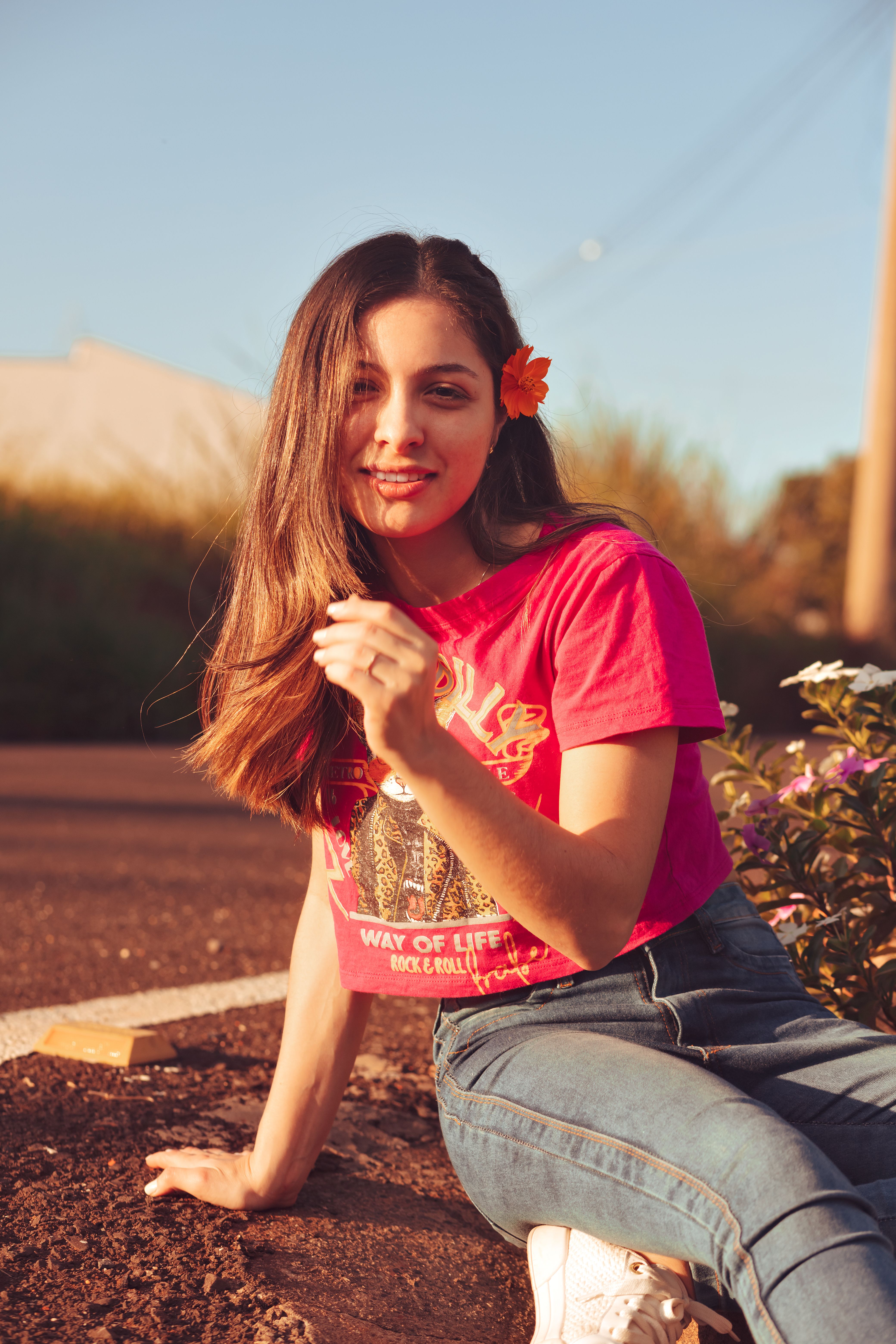 Smiling young woman with a flower in her hair, wearing a red t-shirt and jeans, enjoying the outdoors.