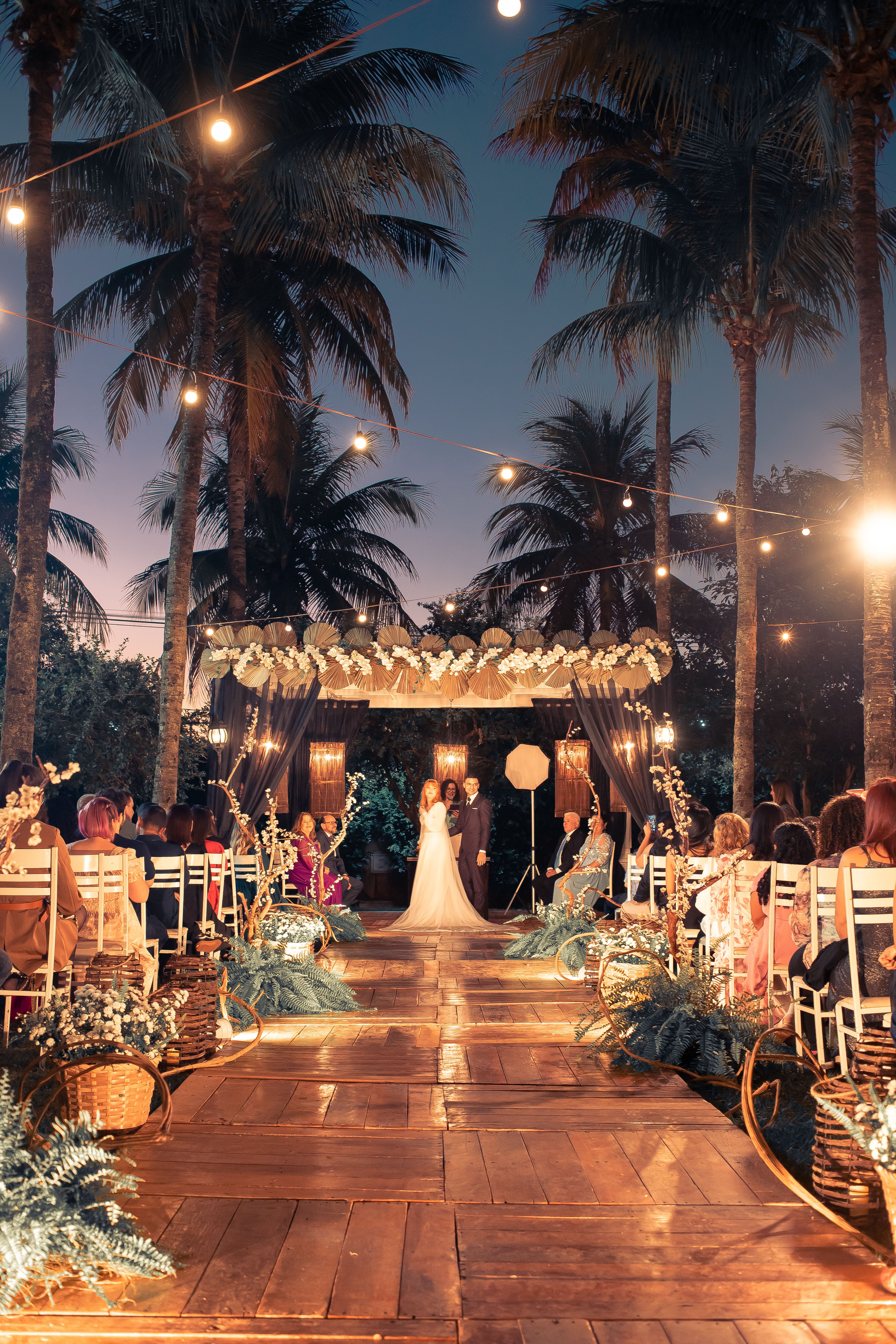A beautiful outdoor wedding ceremony with decorated aisle and elegant lighting under palm trees.