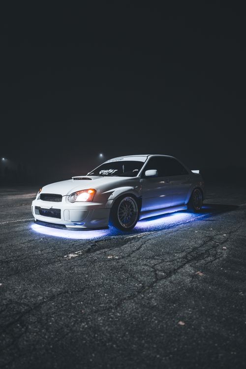 Free A white Subaru car with LED underglow, parked in a dimly lit parking lot at night, showcasing street racing aesthetics. Stock Photo
